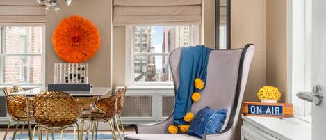 A modern living room with a grey armchair, a dining table with golden chairs, a large orange decorative piece, and a view of the city through large windows.