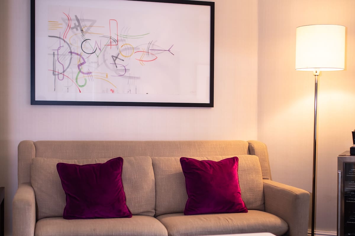 Una habitación de hotel moderna con un sofá color beige, dos cojines morados, una mesa de centro blanca, una lámpara de pie y una obra de arte abstracta enmarcada en la pared.