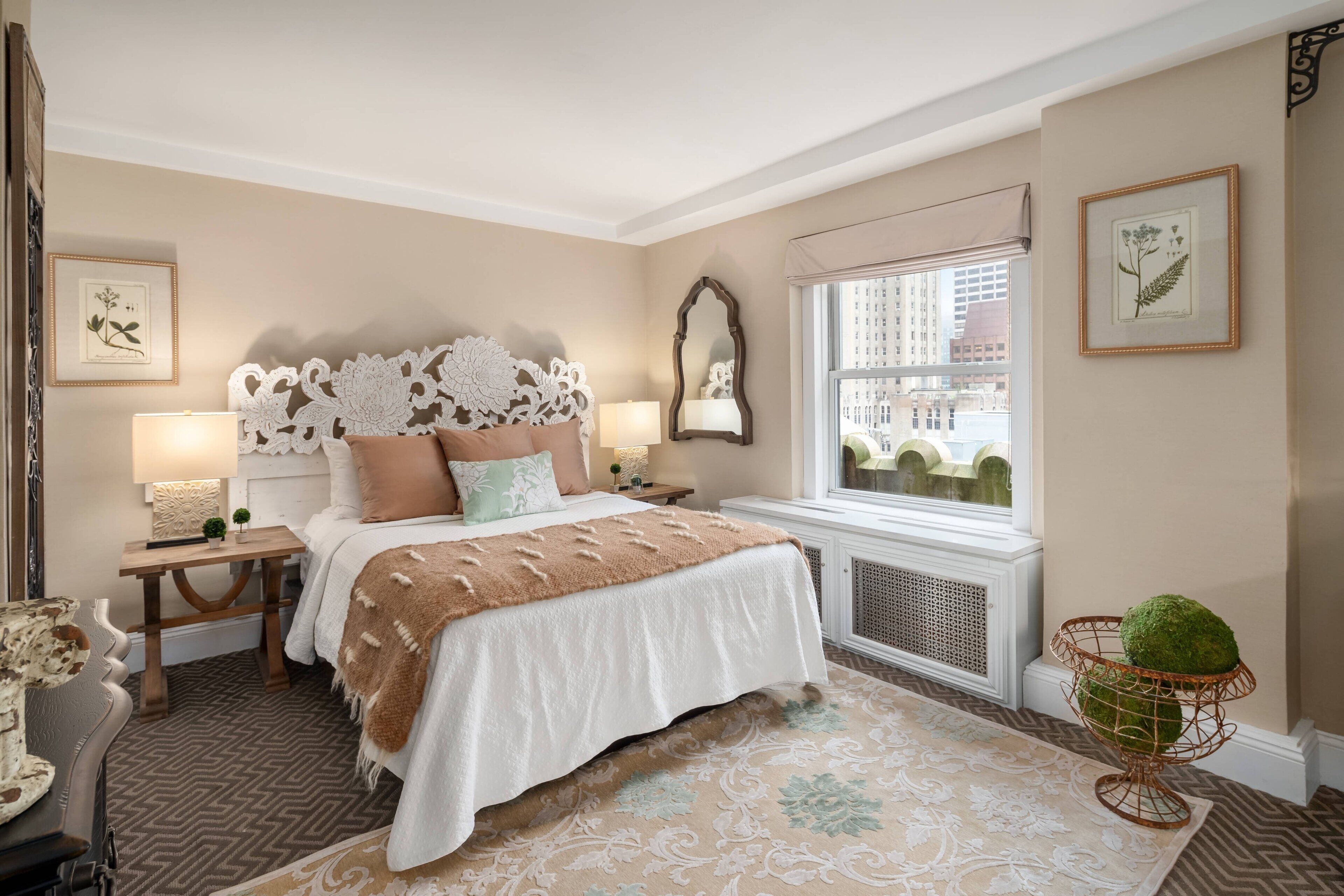 Une chambre dotée d’un grand lit, de deux tables de chevet avec des lampes, d’une fenêtre donnant sur des immeubles et d’œuvres d’art décoratives accrochées au mur.