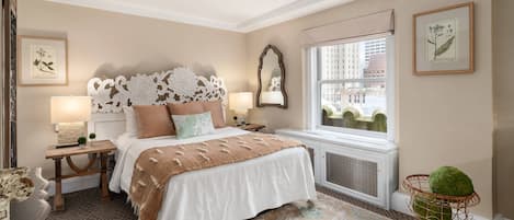Une chambre dotée d’un grand lit, de deux tables de chevet avec des lampes, d’une fenêtre donnant sur des immeubles et d’œuvres d’art décoratives accrochées au mur.