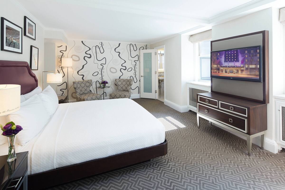 A hotel room with a large bed, a flat-screen TV, a dresser, and a patterned carpet.