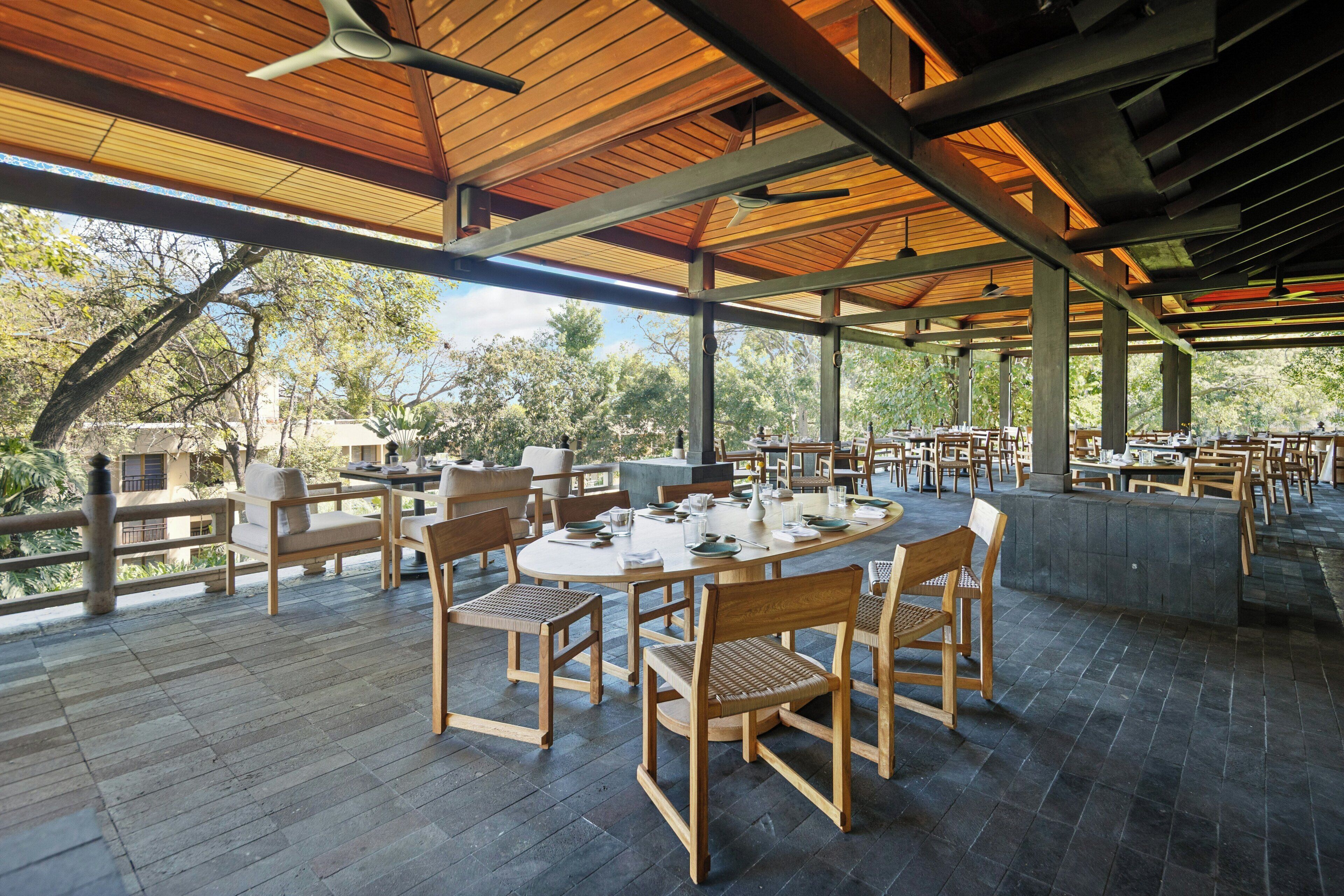 Un espacio de comedor al aire libre con mesas redondas y sillas de madera, rodeado por una terraza de madera y cubierto por una estructura de techo.
