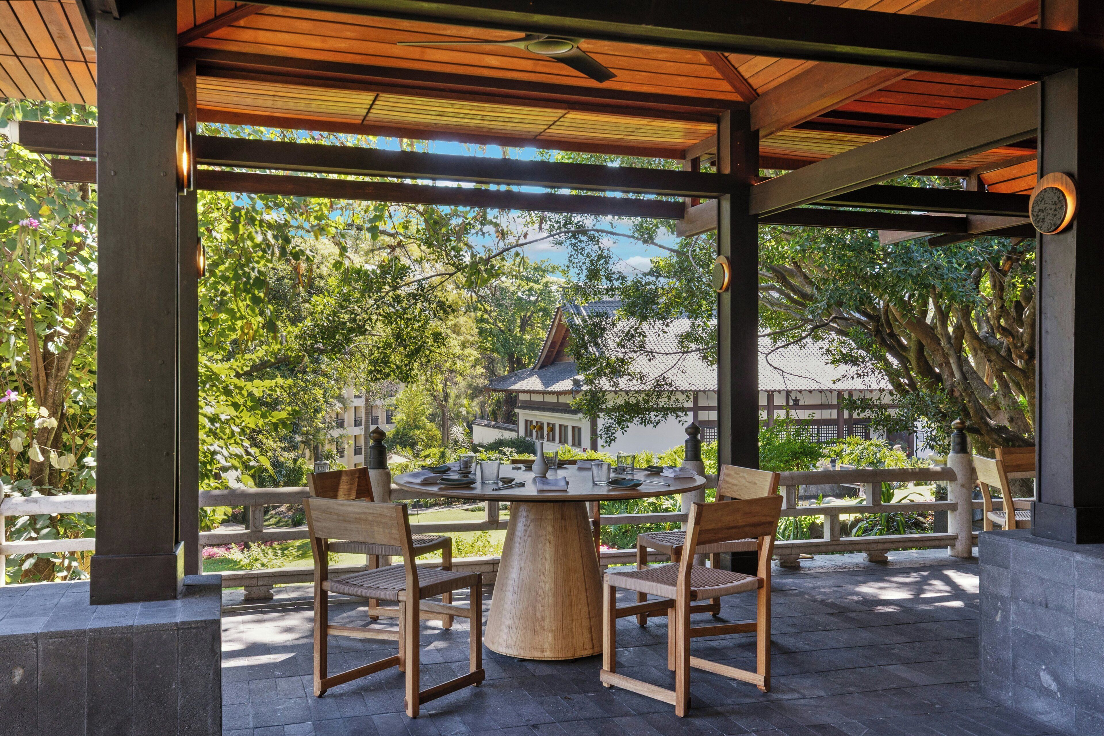 A covered outdoor seating area with a round table and chairs, surrounded by a wooden fence, overlooking a garden and a traditional building.
