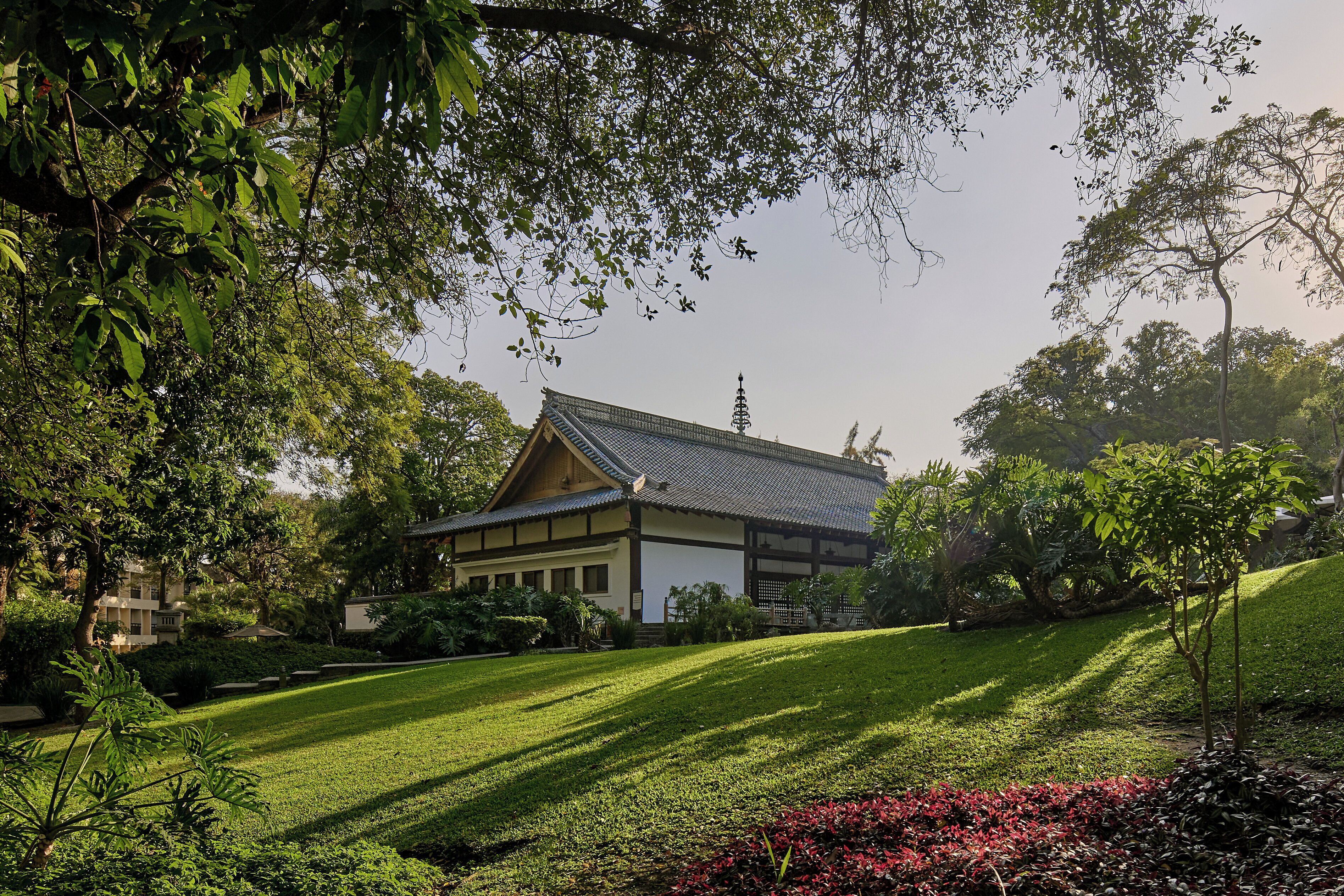Un bâtiment de style japonais traditionnel, doté d’un toit en pente, entouré d’une pelouse bien entretenue et de diverses plantes.