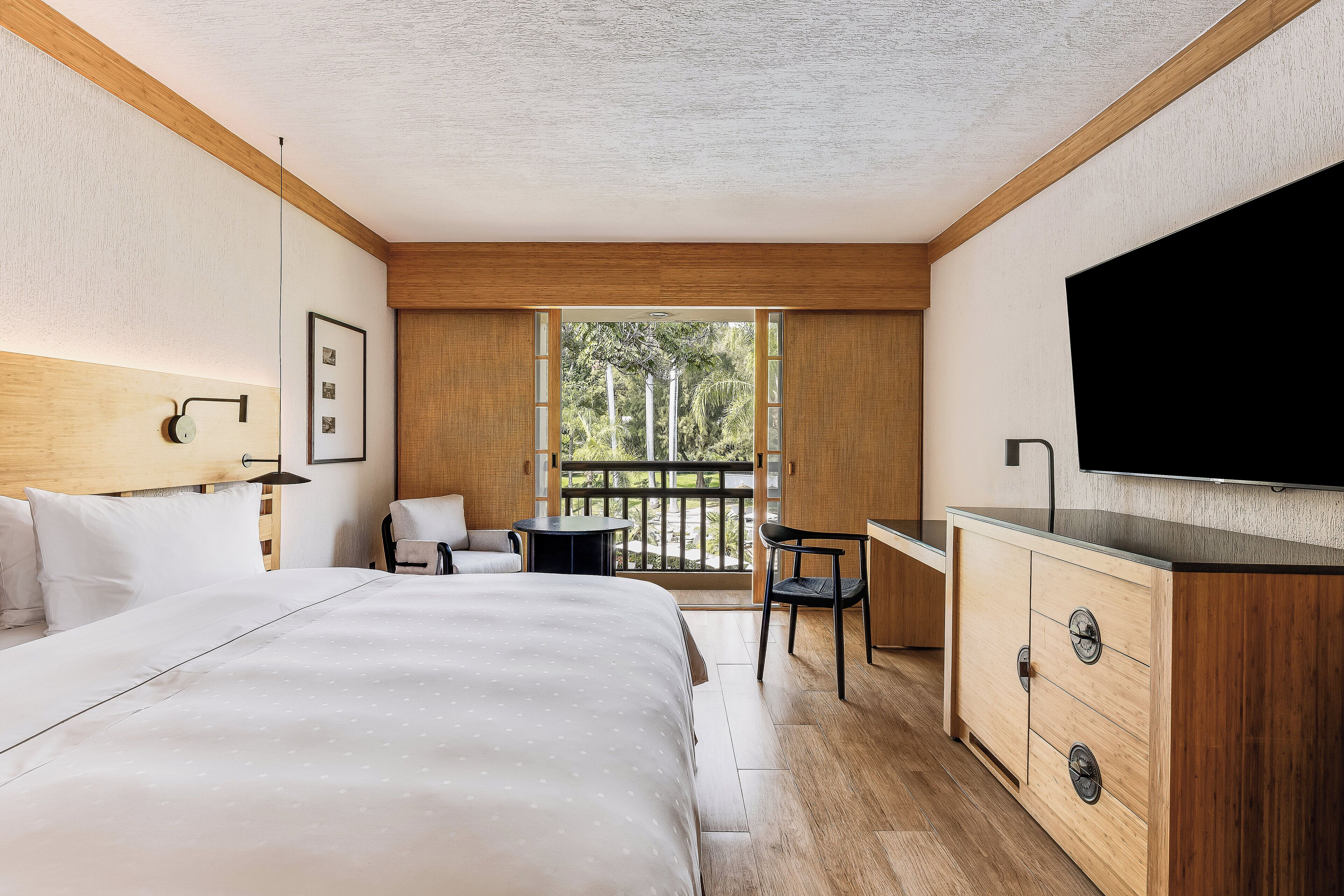 Hotelkamer met een bed, bureau, stoel, televisie en een balkon met uitzicht op bomen.