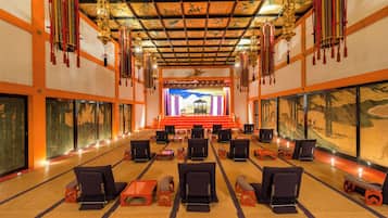 A spacious hall with rows of chairs and tables, a stage at the front, and traditional wall paintings.