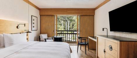 Habitación de hotel con una cama grande, un escritorio, una silla, un televisor y un balcón con vistas a árboles.