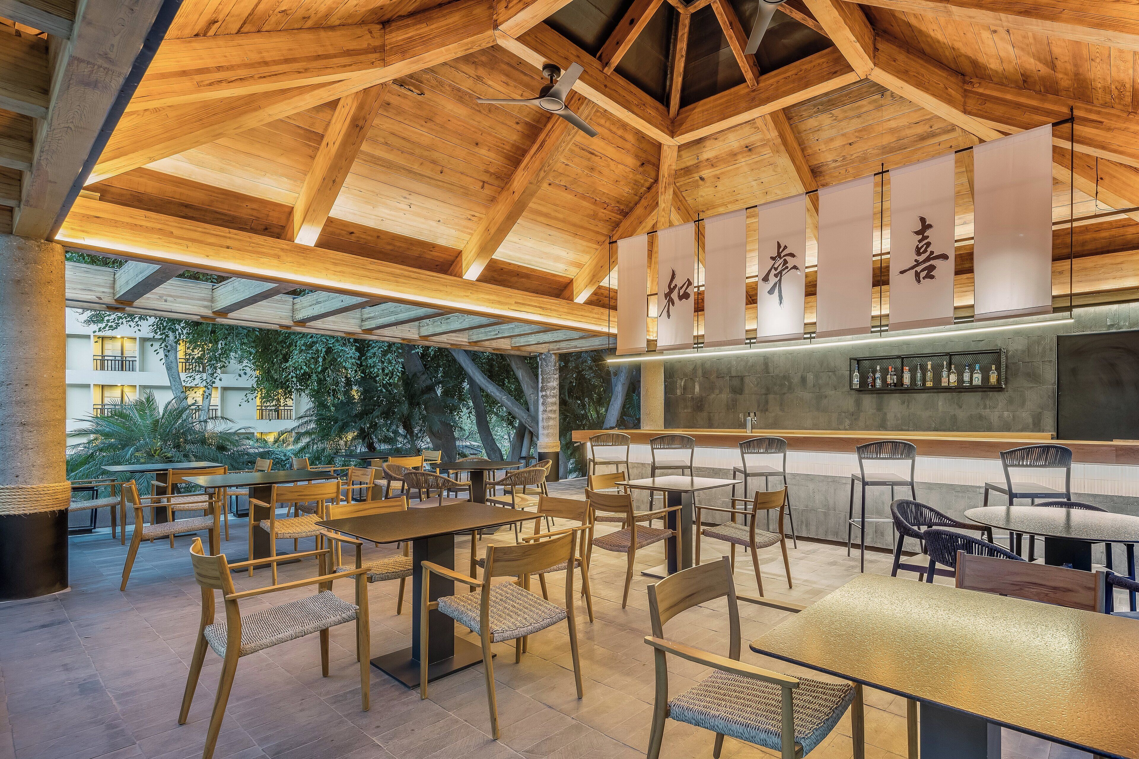 A spacious outdoor dining area with wooden tables and chairs, a bar with bottles, and a wooden ceiling structure.