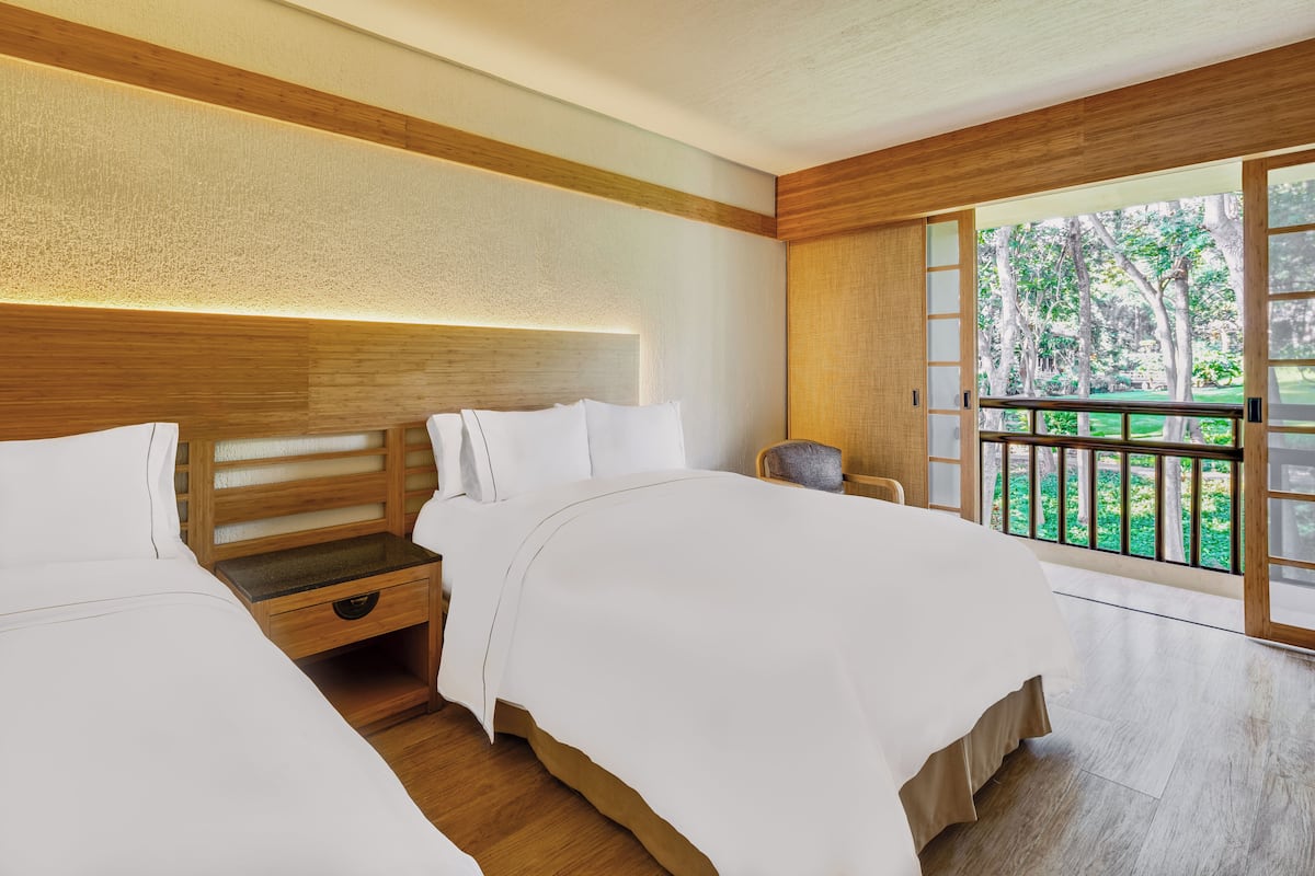 A hotel room with two beds, a wooden headboard, a nightstand, and a view of a balcony with trees.
