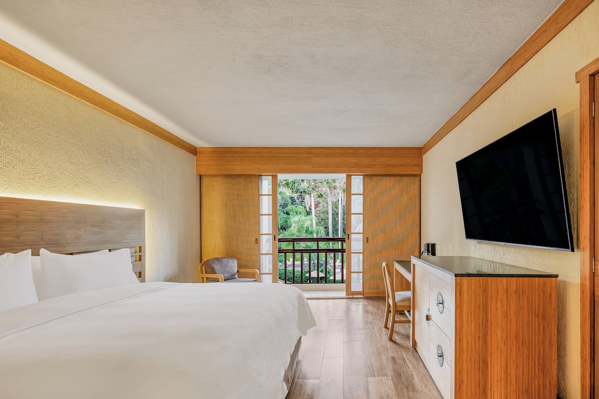 A hotel room with a large bed, a desk, a TV, and a balcony with a view of greenery.