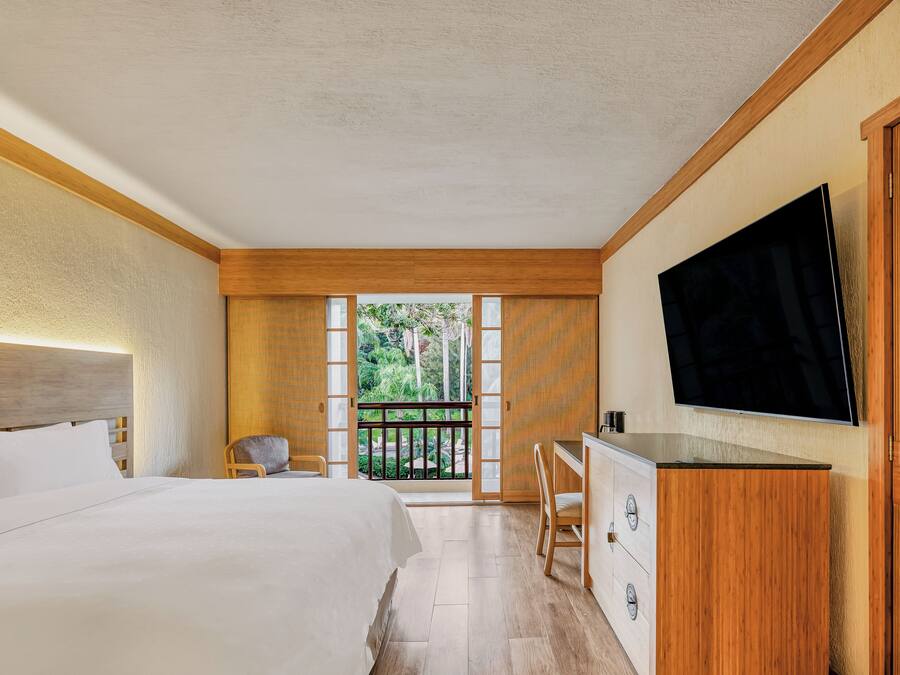 Habitación de hotel con una cama grande, un escritorio, televisión y balcón con vistas a zonas verdes.