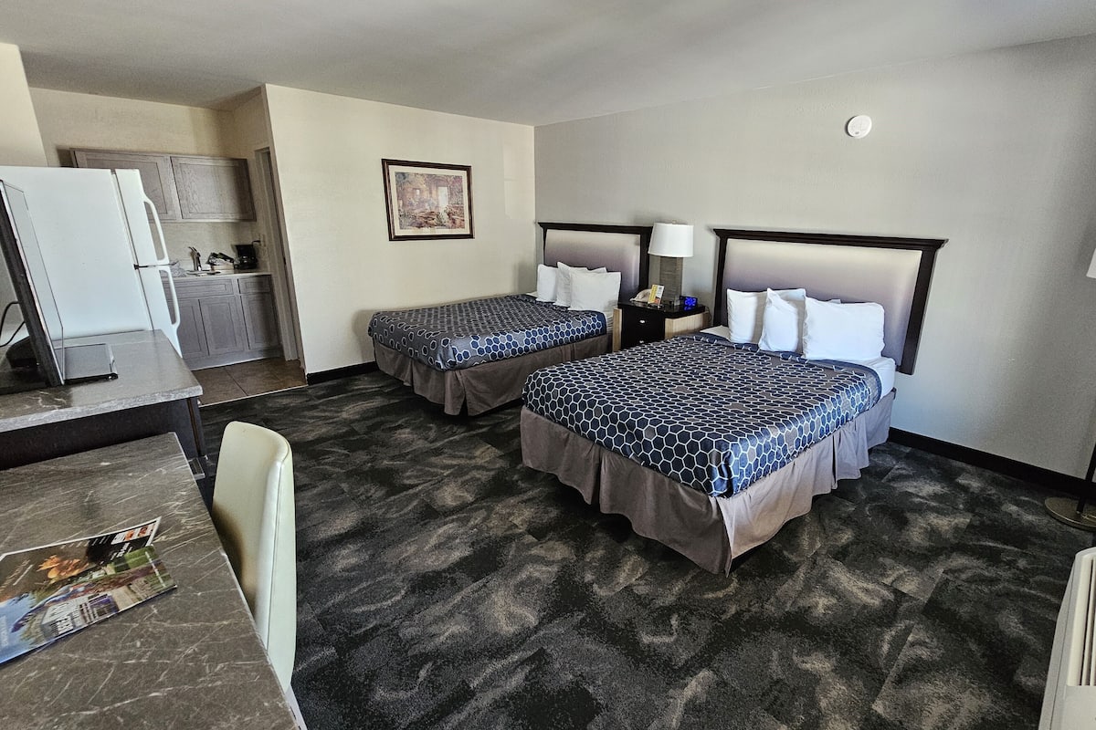 A hotel room with two beds, a desk, a chair, a small kitchenette, and a framed picture on the wall.