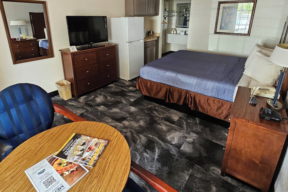 A hotel room with a bed, a TV, a refrigerator, a small kitchenette, a mirror, and a round table with a magazine.