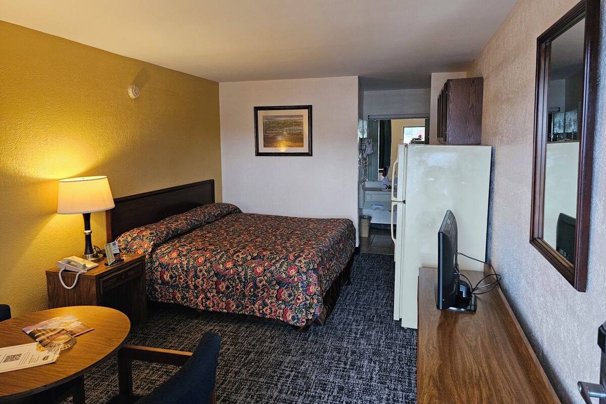 A hotel room with a bed, a desk with a lamp, a refrigerator, and a bathroom visible through an open door.