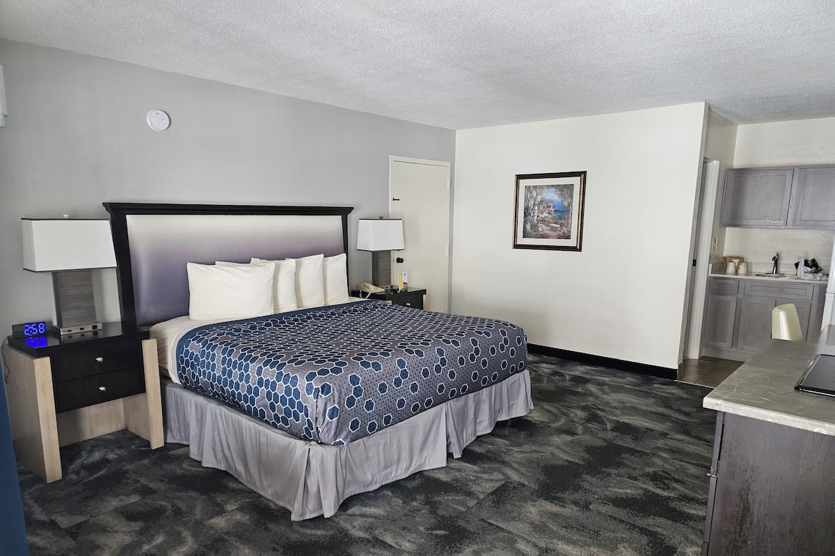 A hotel room with a bed, nightstands, a desk, a chair, a painting, and a kitchenette.