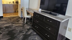 A compact living space with a kitchenette, a dark wood desk with drawers, a flat-screen TV, and a patterned carpet.