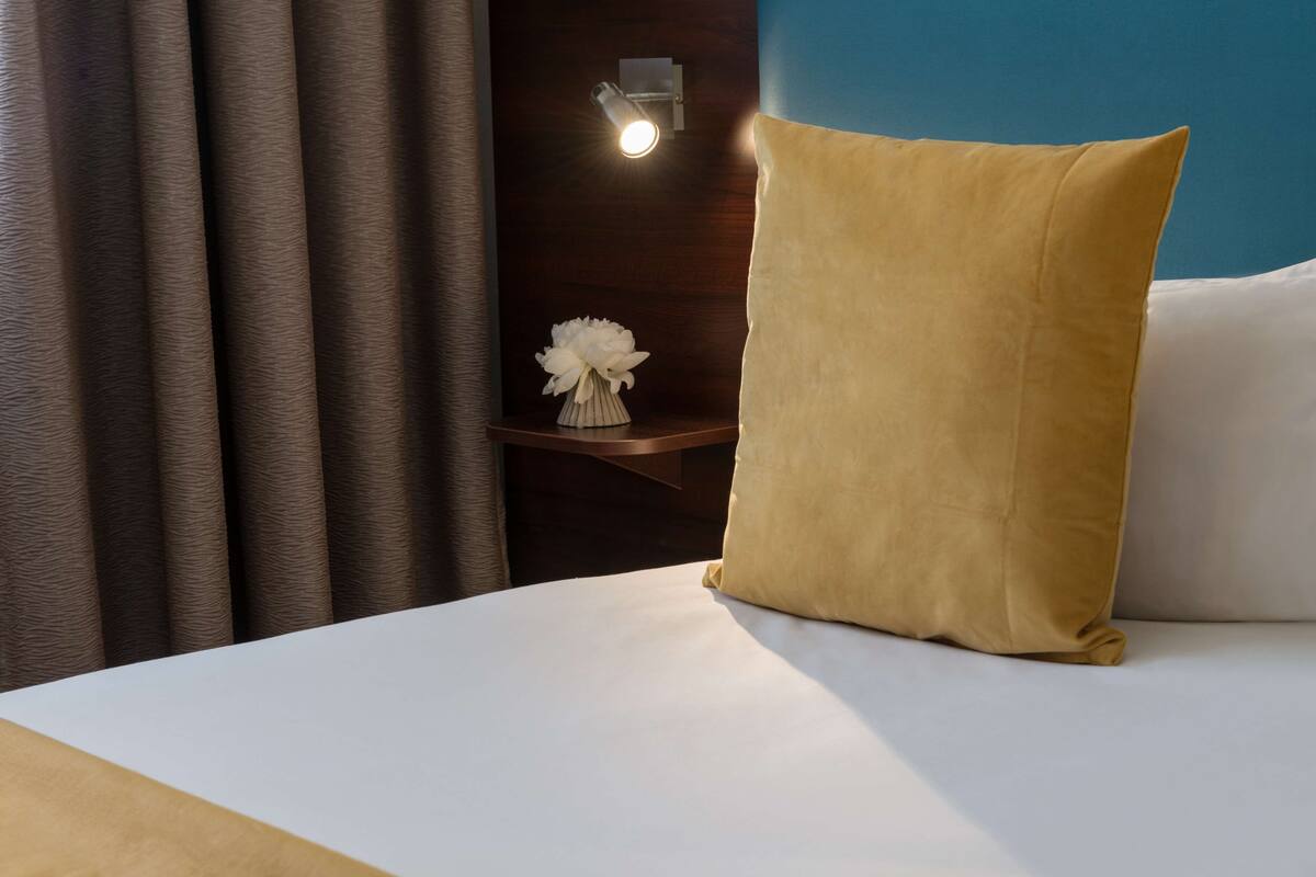 Una cama bien tendida con un único y grande almohadón, una mesita de noche con un florero y una lámpara de pared.