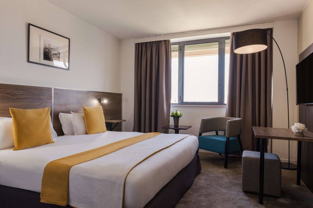 Habitación de hotel con una cama grande, un escritorio, una silla, una mesita y una ventana con cortinas.