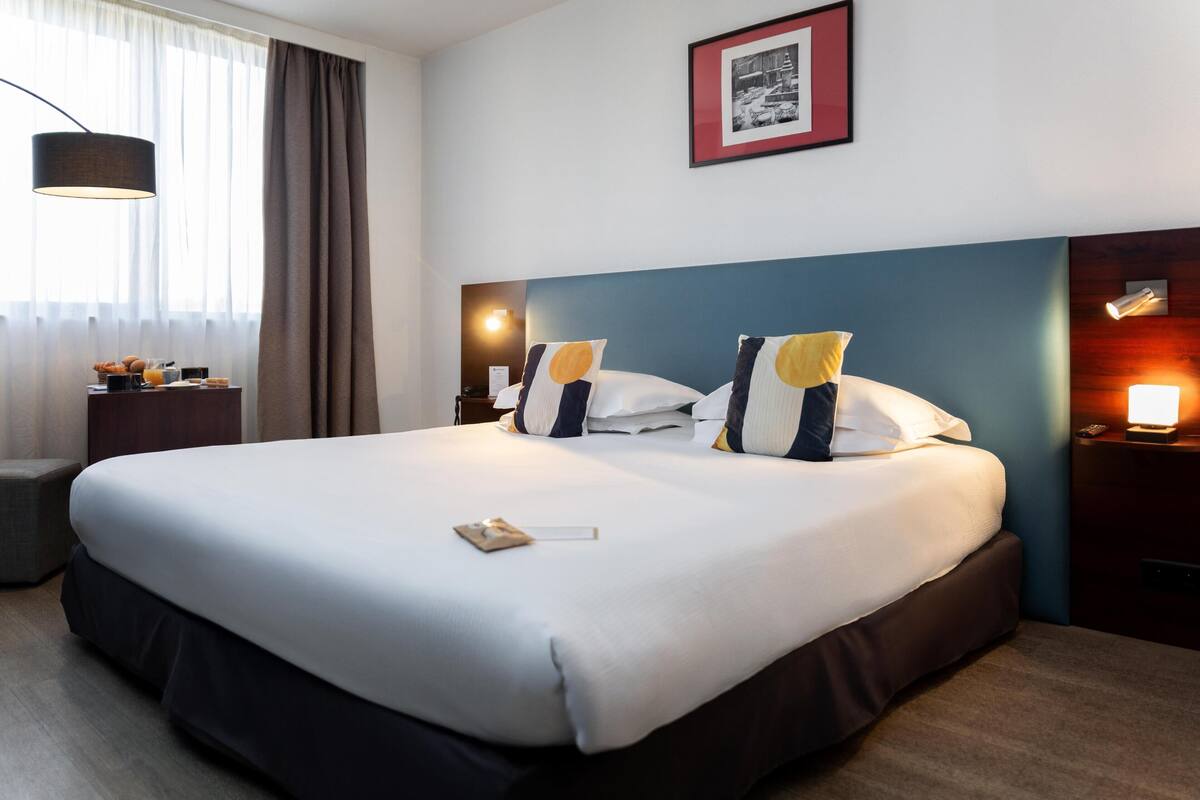 Habitación de hotel con una cama grande, mesitas de noche, una lámpara, un cuadro en la pared y una ventana con cortinas.