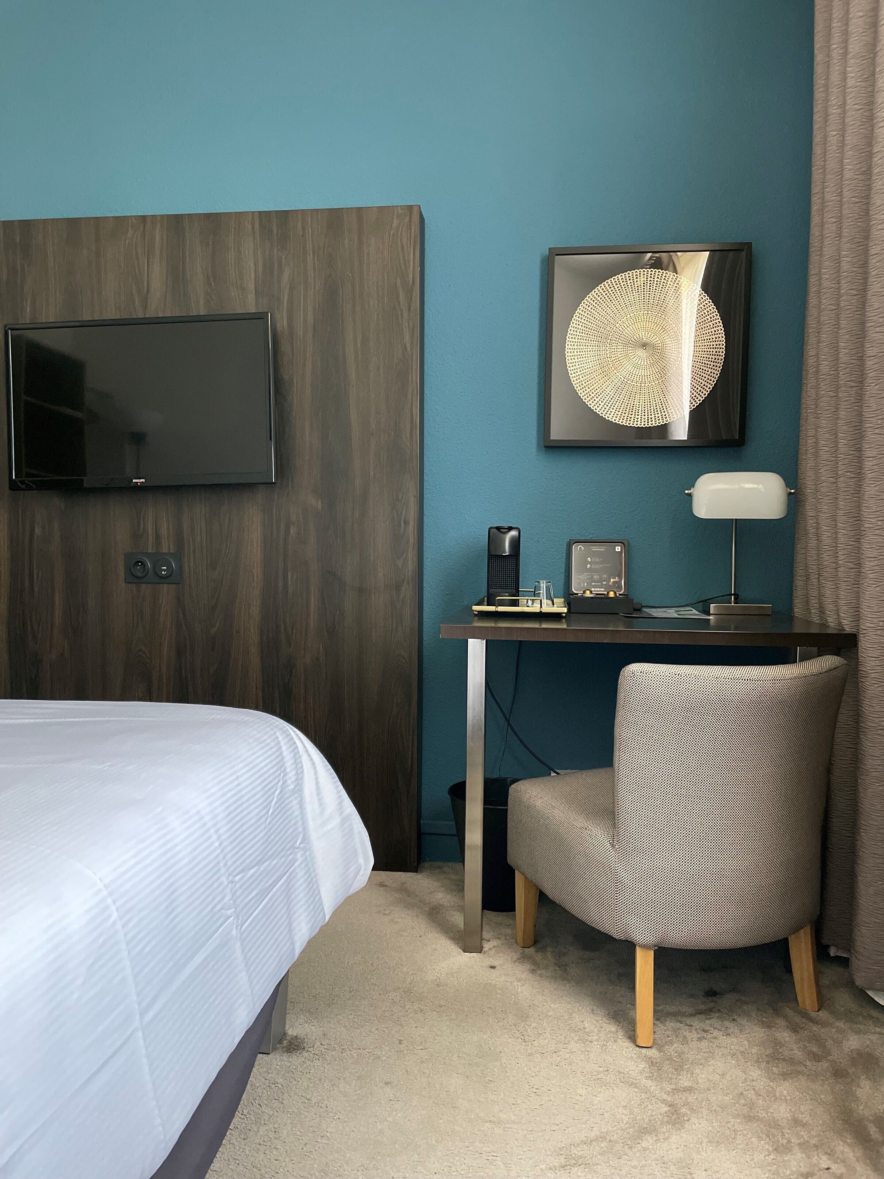 A hotel room with a bed, a desk with a chair, a flat-screen TV, a decorative wall piece, and a lamp.