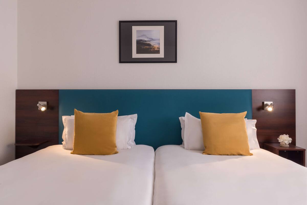 Una cama doble con almohadas blancas y mostaza, un cabecero verde azulado y un cuadro enmarcado encima.