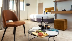Una habitación de hotel con una cama, una mesa de comedor con comida, una silla, una botella de vino y una mesita de noche con una lámpara.