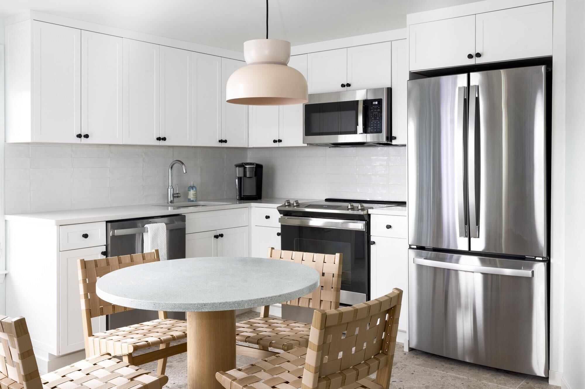 A modern kitchen with white cabinets, stainless steel appliances, a round table, and woven chairs.