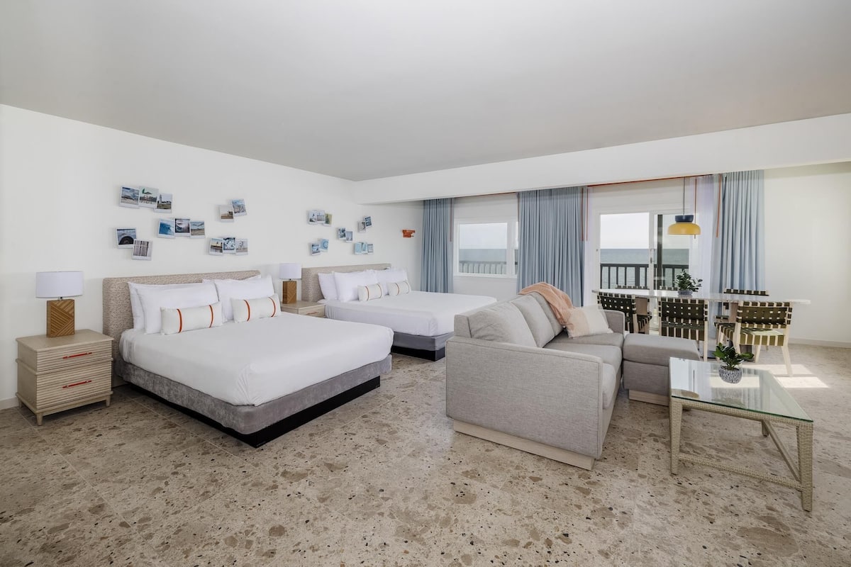 A hotel room with two beds, a sofa, a small table, and a balcony with a view.