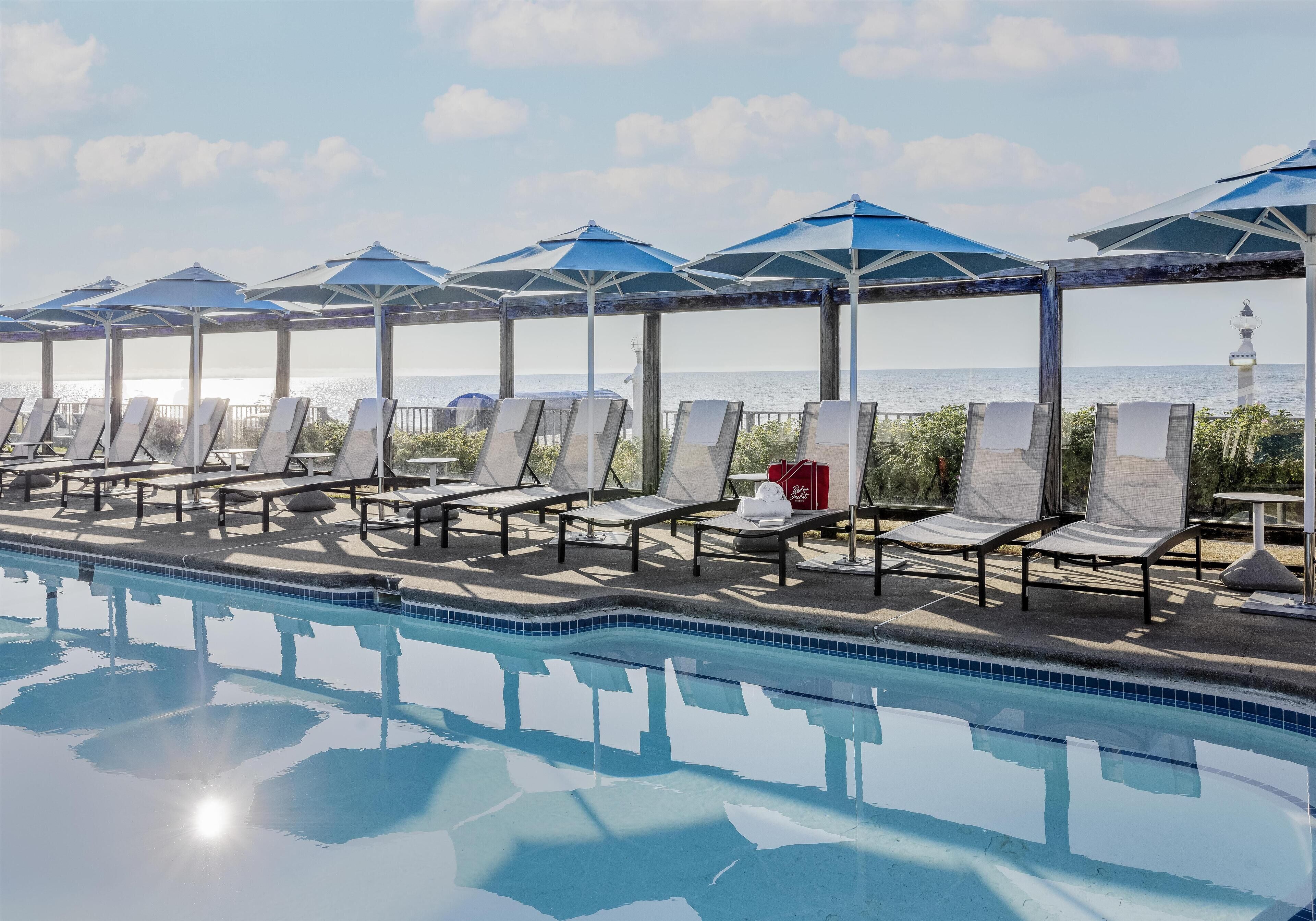 A swimming pool with lounge chairs and umbrellas, a view of the sea, and a lighthouse in the background.
