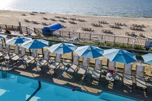 Een zwembadgedeelte met ligbedden en parasols, een strand met ligstoelen en een helder uitzicht op de oceaan.