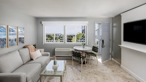 A modern living room with a grey sofa, a glass coffee table, a dining area with chairs, a large window with a view, and a mounted TV. - Blue Water Resort (South Yarmouth)