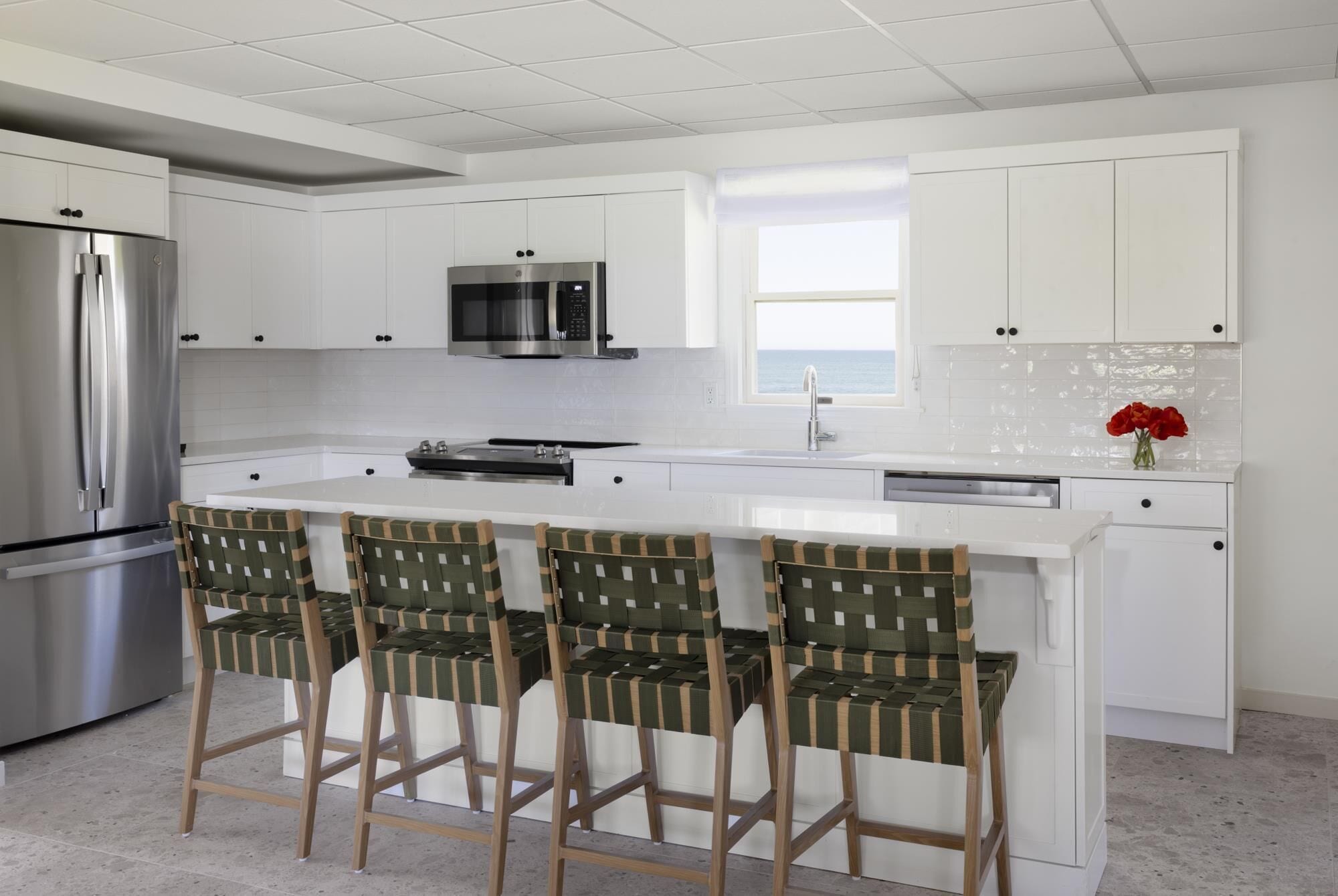 Een moderne keuken met witte kasten, een roestvrijstalen koelkast, een magnetron en een vaatwasser. Er staan drie geweven stoelen bij het eiland.