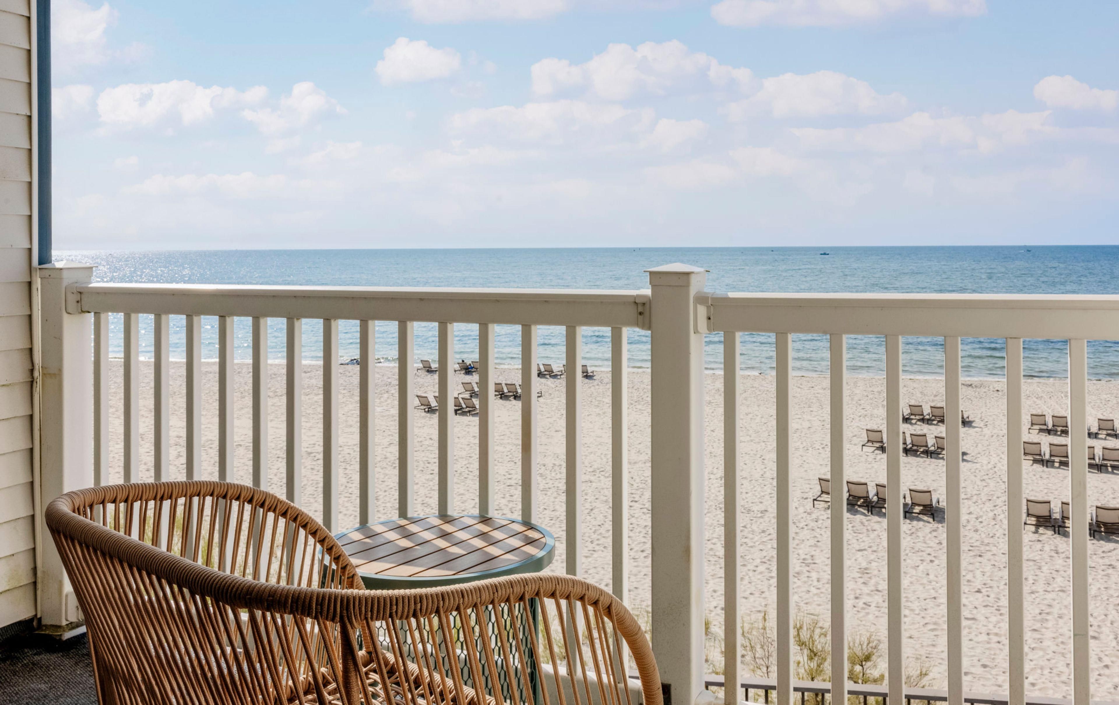 A balcony with wicker chairs and a table overlooking a sandy beach with lounge chairs.