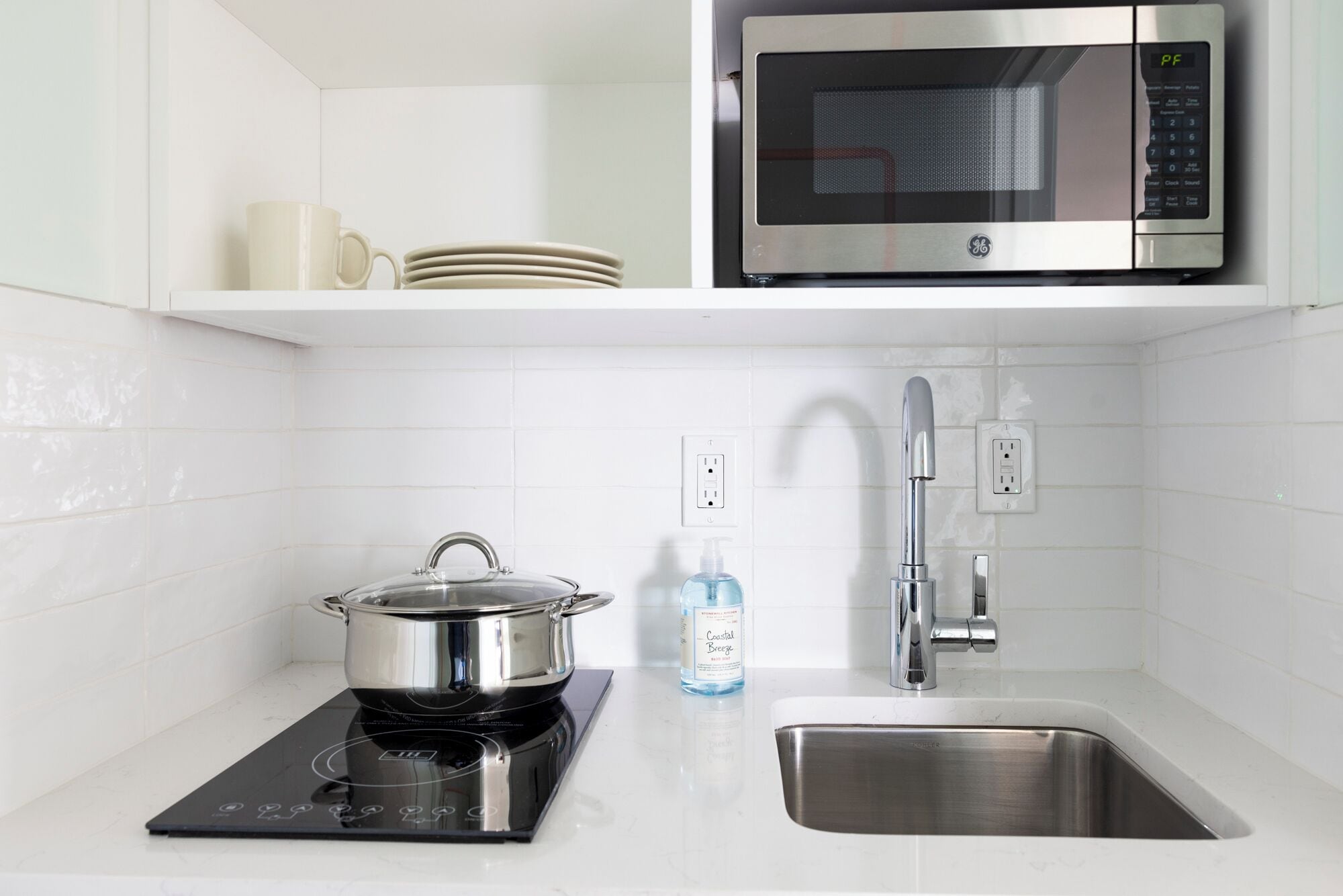 A kitchen with a black induction cooktop, a stainless steel sink, a microwave, and a bottle of hand soap.