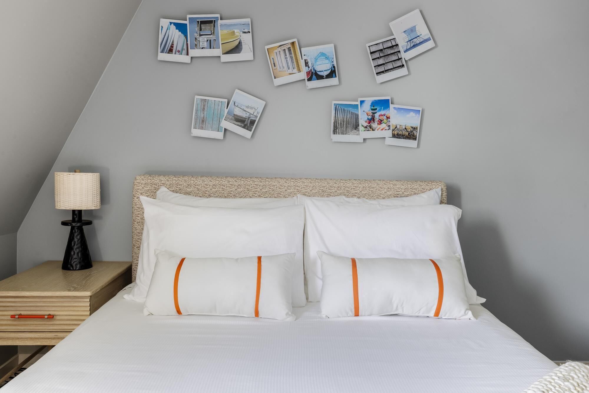 A neatly made bed with white bedding and orange-striped pillows, a bedside table with a lamp, and a wall adorned with various photographs.