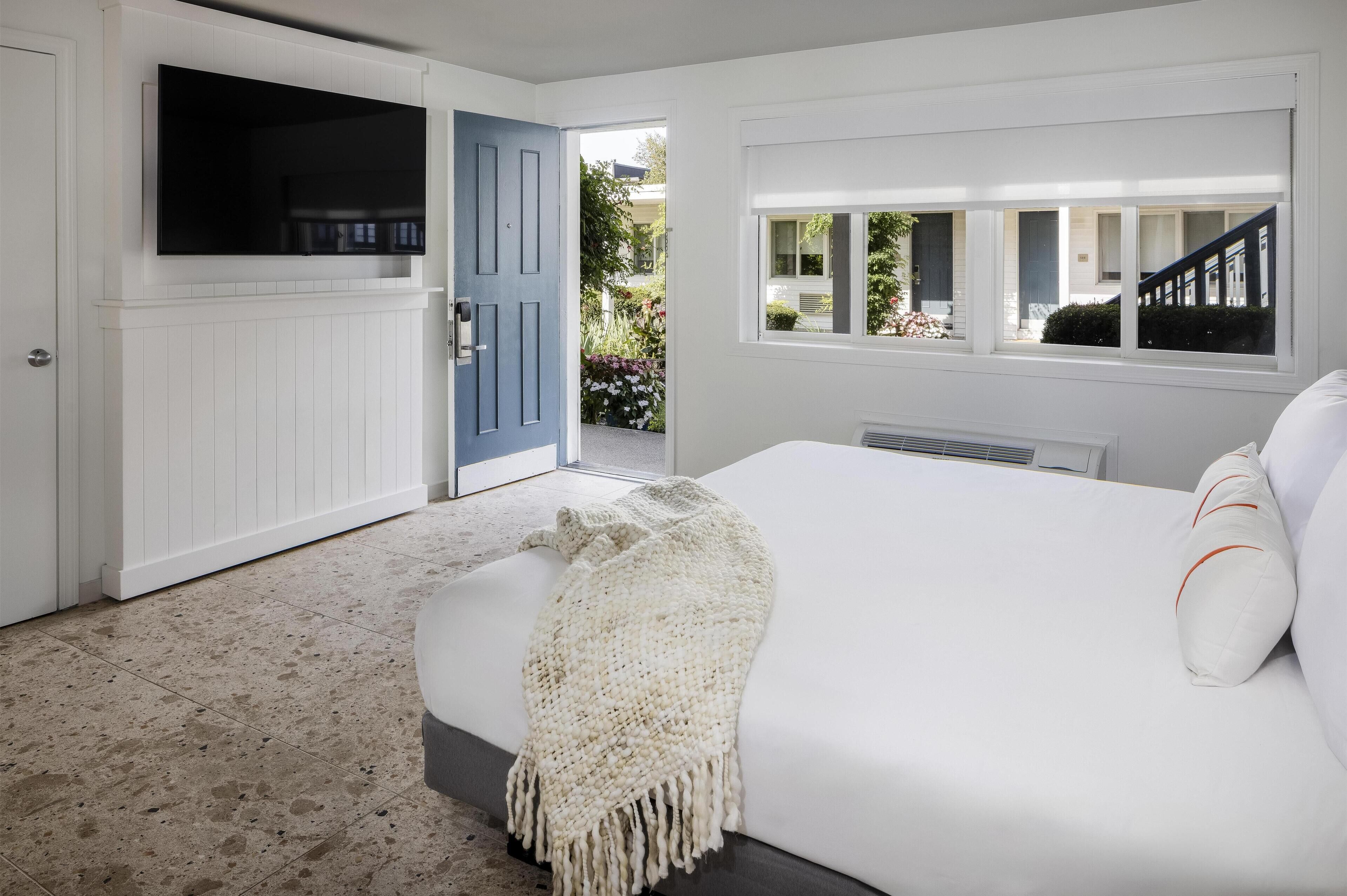 A bedroom with a large bed, a TV mounted on the wall, a door leading outside, and a window with a view of the outside.