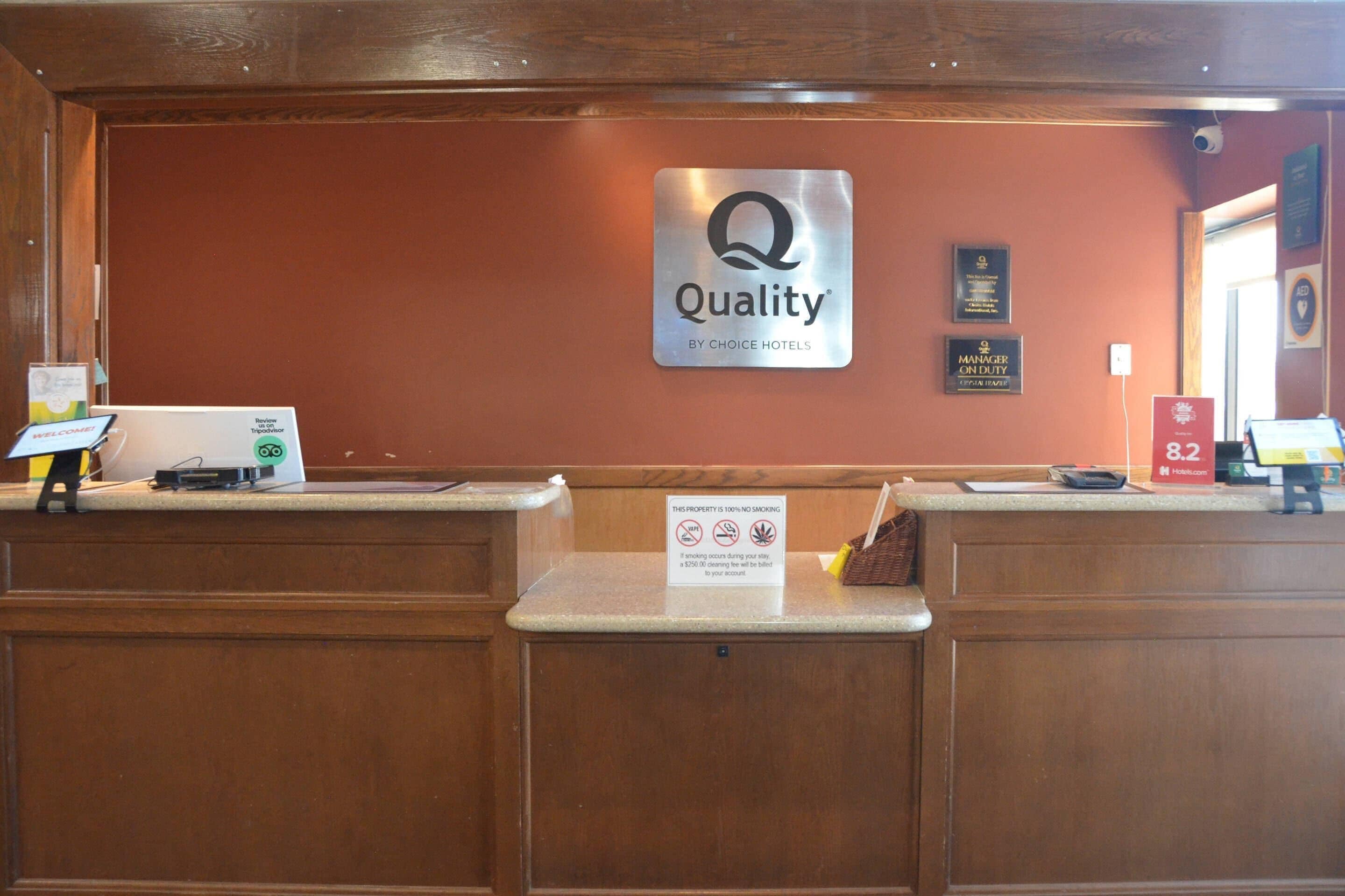 Um balcão de recepção com a placa 'Quality by Choice Hotels', uma placa 'Gerente de Plantão' e uma placa 'Proibido Fumar'.