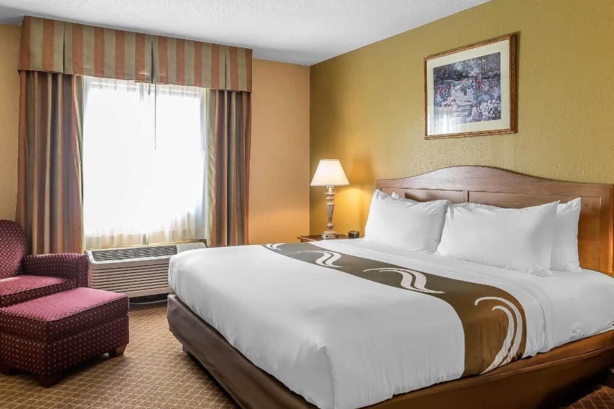 Quarto de hotel com uma cama grande, uma cadeira, um abajur e um quadro na parede.