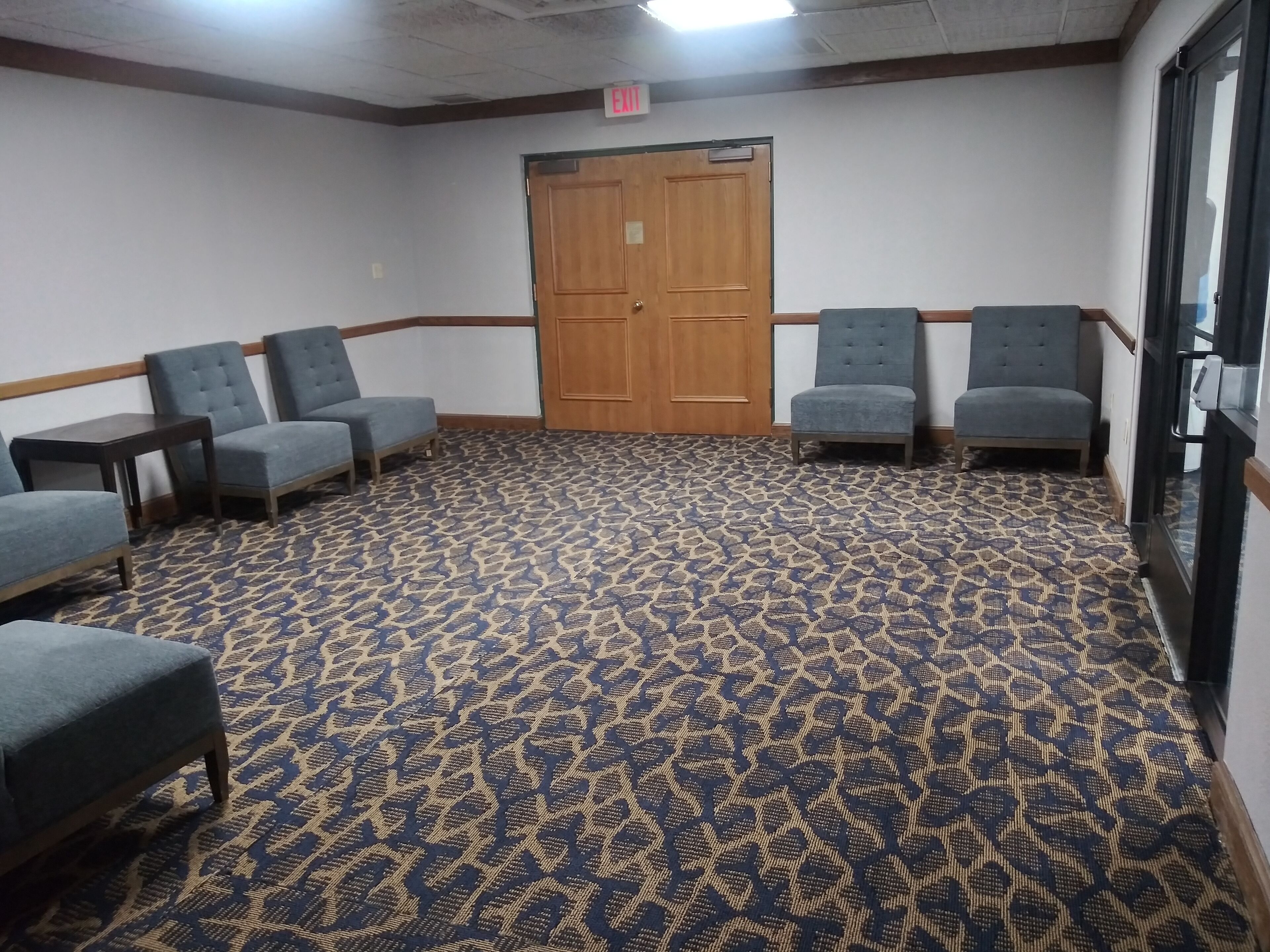 A room with a patterned carpet, four grey armchairs, a wooden door, and a glass door.