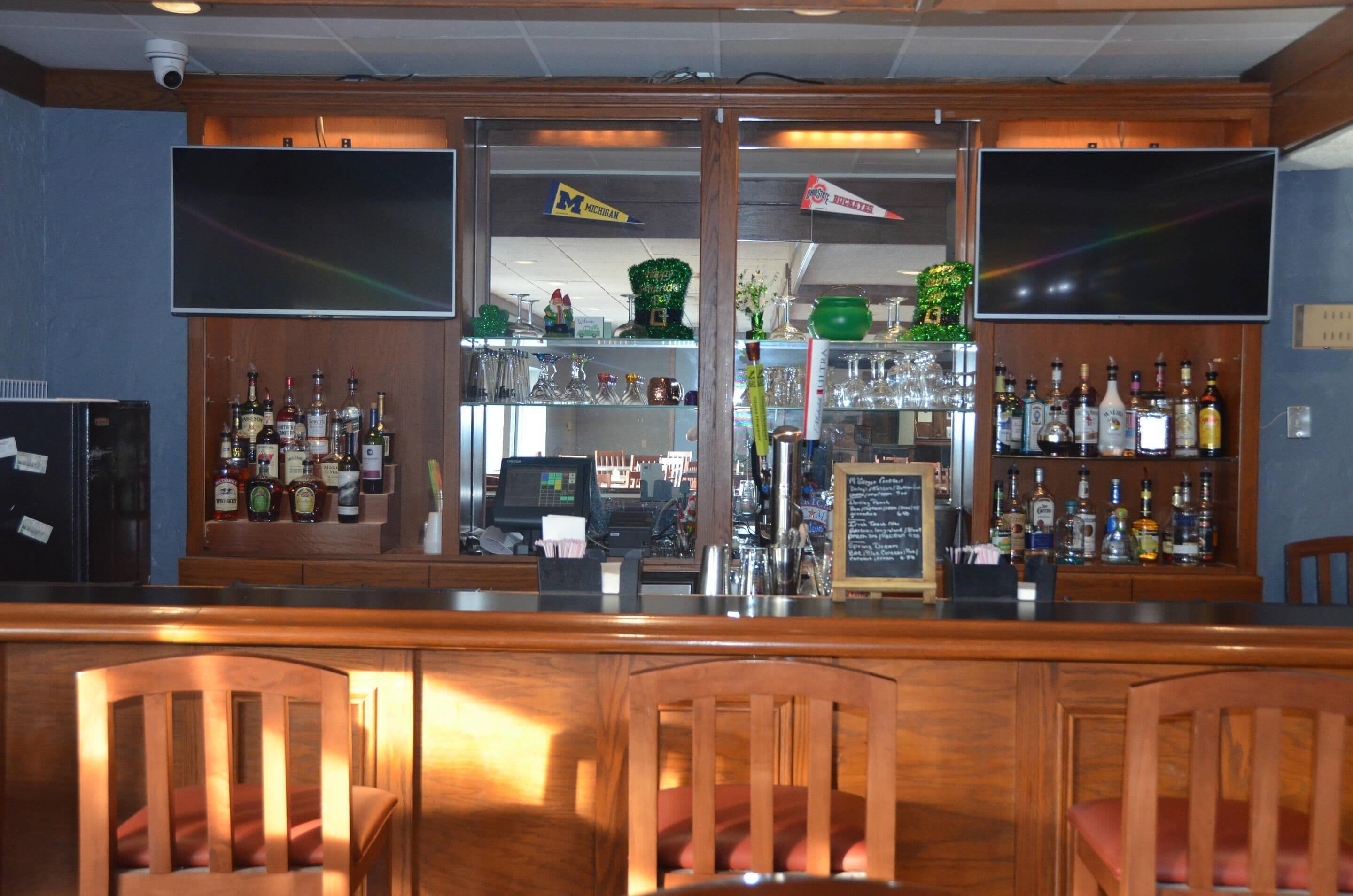 Zona de bar con mostrador de madera, varias botellas de licor, una caja registradora y un televisor de pantalla plana grande montado en la pared.