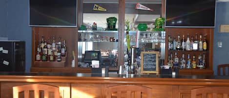 Zona de bar con mostrador de madera, varias botellas de licor, una caja registradora y un televisor de pantalla plana grande montado en la pared.