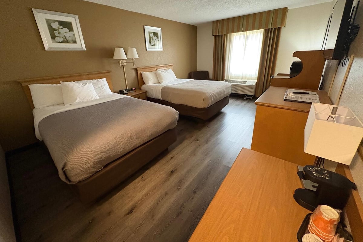 Quarto de hotel com duas camas, uma escrivaninha, uma cadeira, uma televisão e uma janela com cortinas.