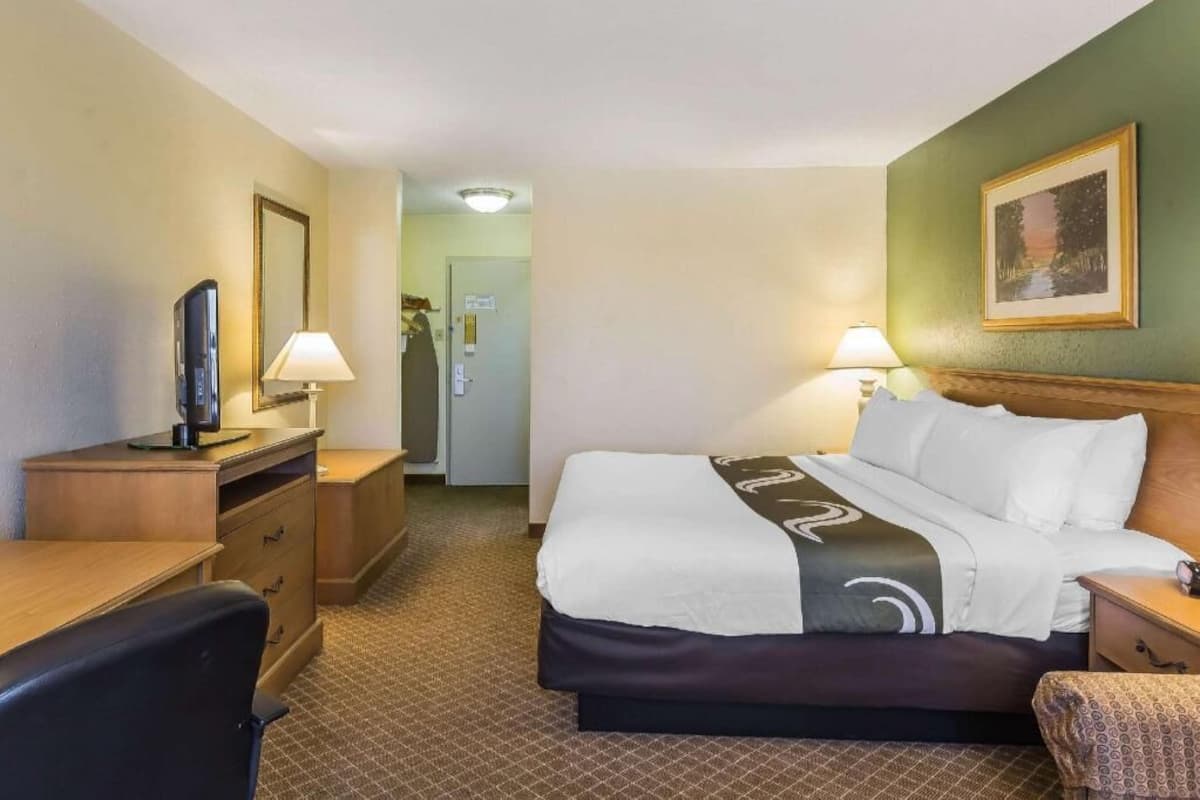 Quarto de hotel com cama, escrivaninha, cadeira, luminárias e um quadro na parede.