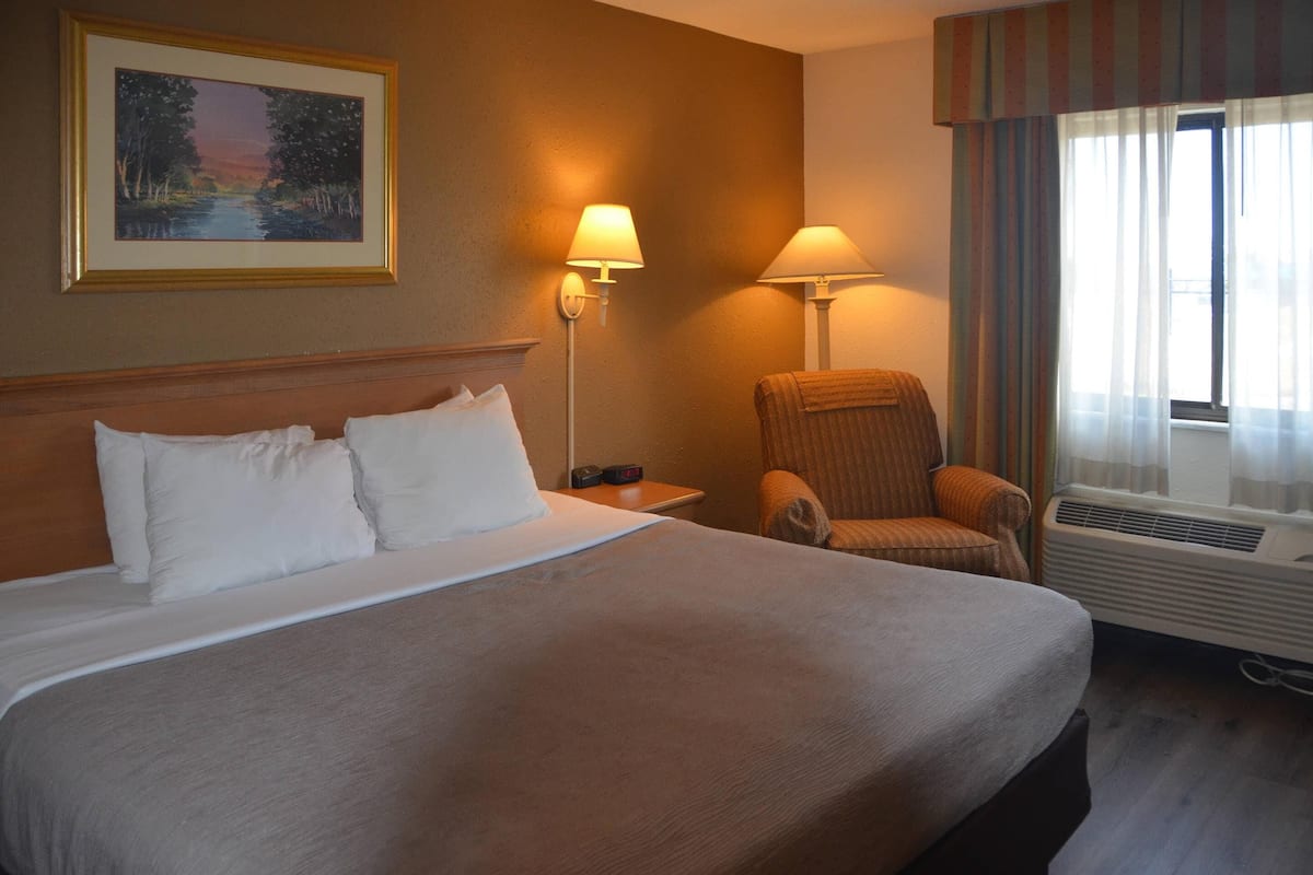 Quarto de hotel com uma cama grande, duas luminárias de cabeceira, uma cadeira, uma janela com cortinas e um quadro na parede.