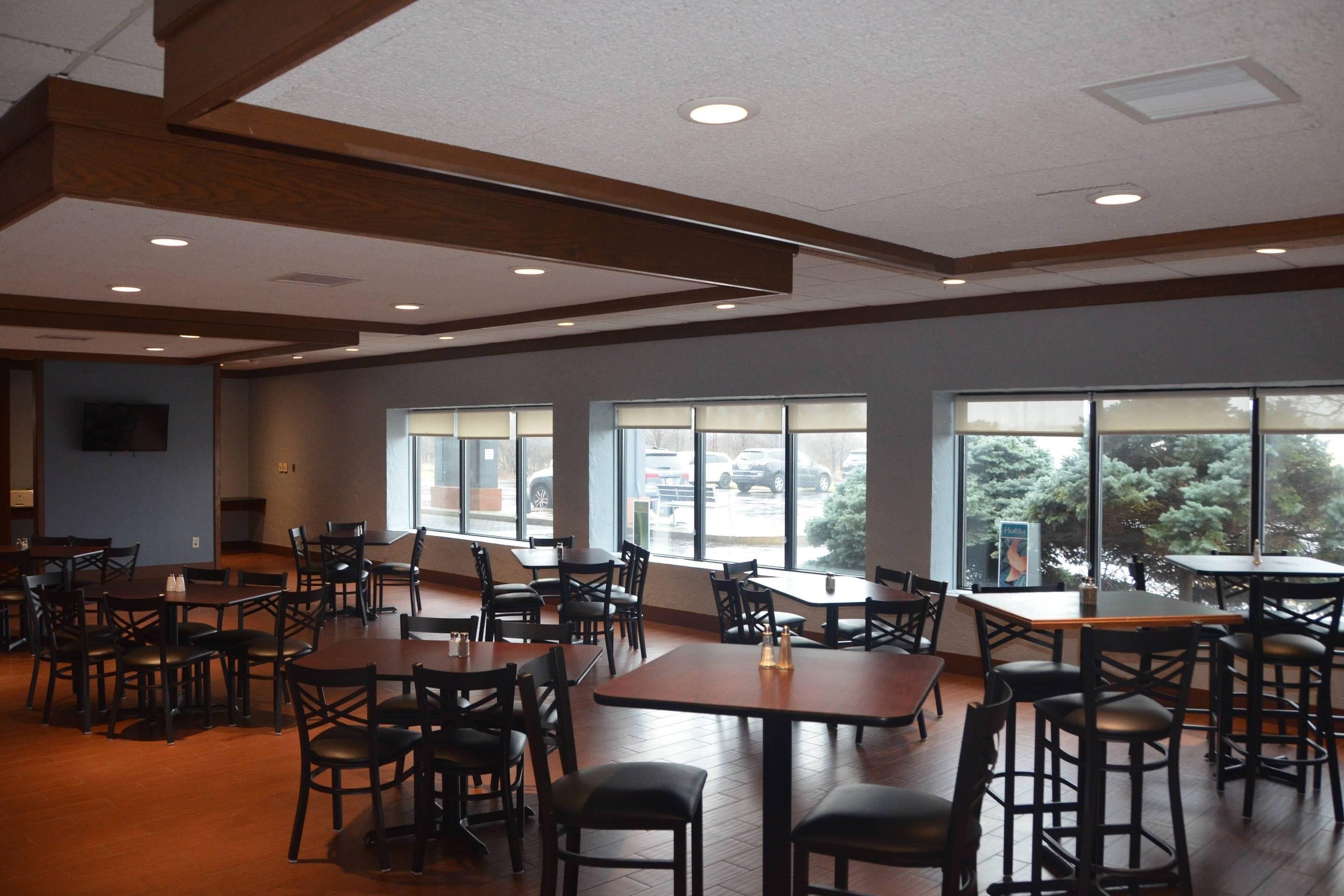 A spacious indoor dining area with multiple tables and chairs, large windows with blinds, and a wooden ceiling structure.