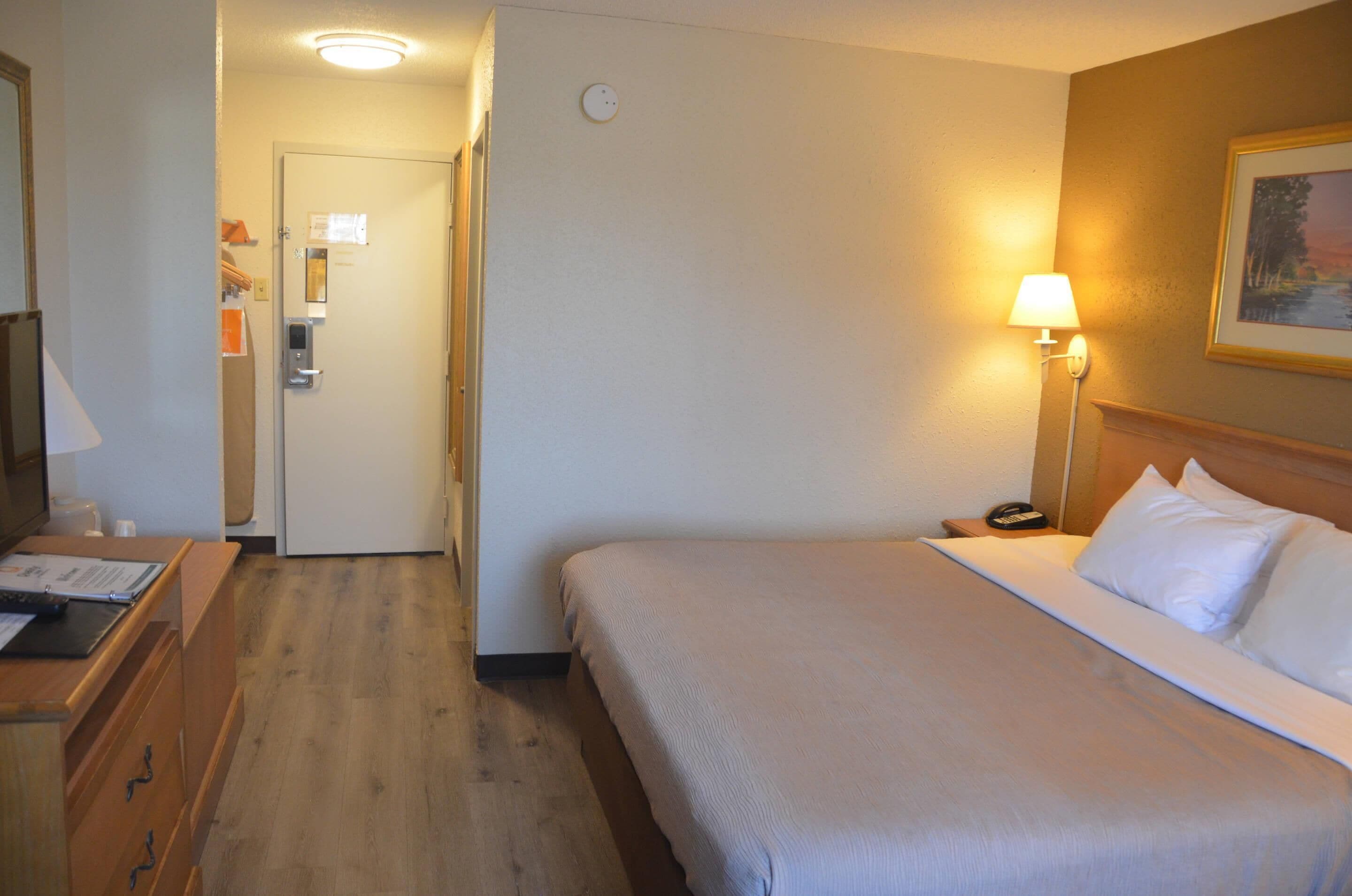 Habitación de hotel con cama, mesita de noche, lámpara y una puerta que da a otra habitación.