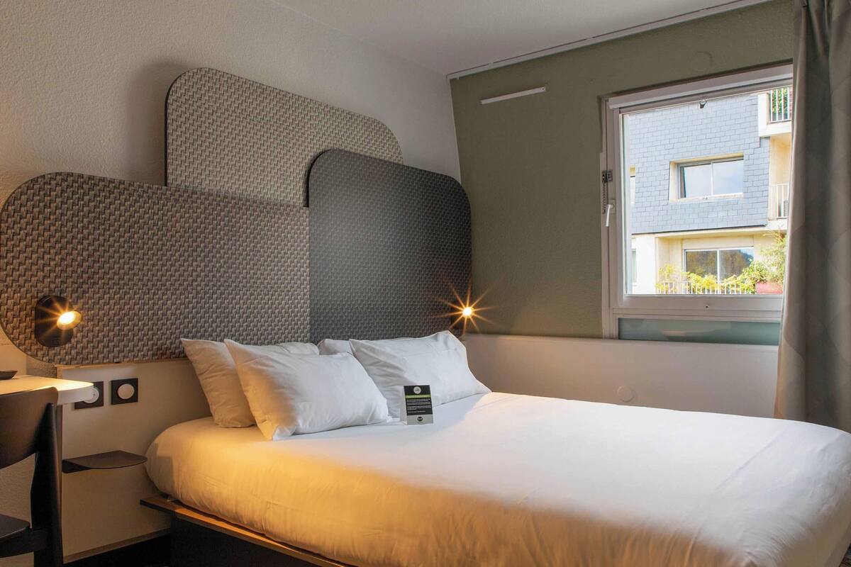 A hotel room with a bed, a desk, and a window with a view of a building.