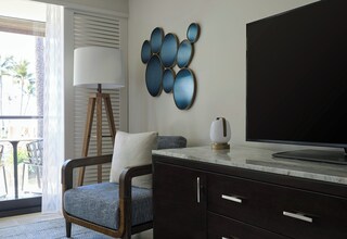 A modern living room with a dark wooden cabinet, a large flat-screen TV, a blue upholstered chair, a white floor lamp, and a wall art piece.