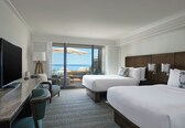 A hotel room with two beds, a desk with a TV, and a view of the beach.