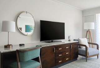 A modern hotel room with a flat-screen TV, a round mirror, a desk with a lamp, a chair, and a large window.